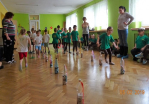 Dzieci stoją z nauczycielkami w rzędach. Na podłodze rozstawione są plastikowe butelki. Dzieci okrążają je slalomem.